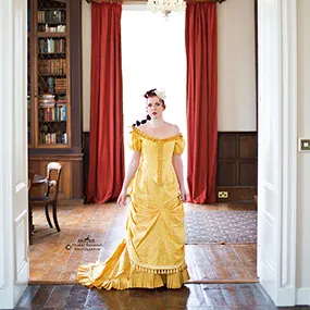 A redhaired girl wearing a yellow Victorian ballgown in a Queen Anne manor house